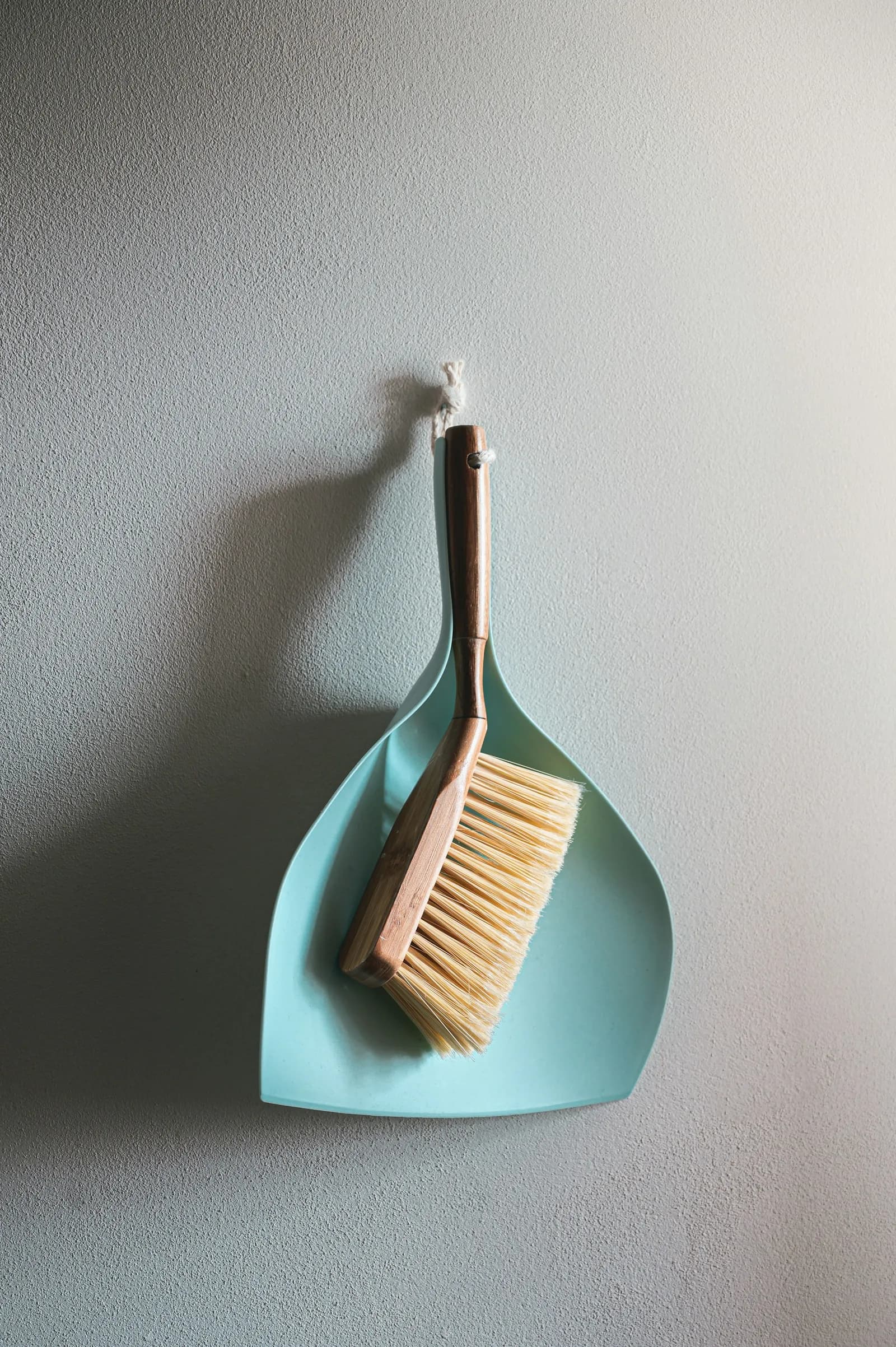 A small brush and a blue dustpan hang on a white wall