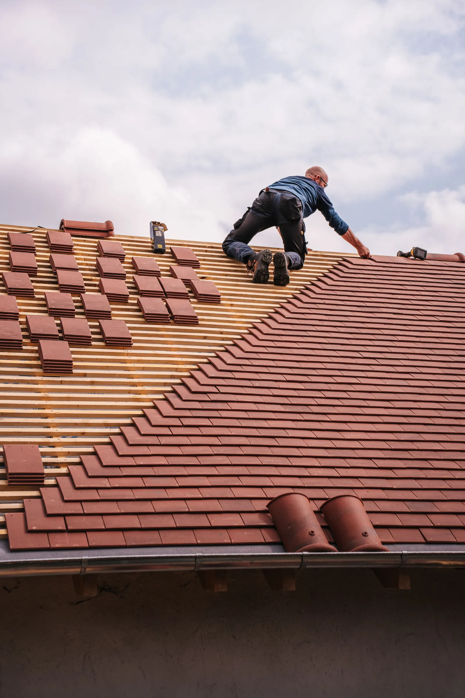 A roofer kneels on a roof and lays rows of clay tile